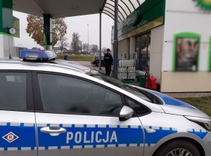 na pierwszym planie widać policyjny oznakowany radiowóz zaparkowany przed stacją paliw. Umundurowana policjantka idzie w kierunku drzwi wejściowych na stację