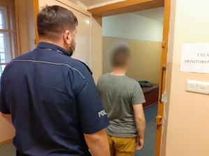 w drzwiach otwartej celi stoi zwrócony tyłem do obiektywu mężczyzna. Za nim stoi policjant służby dyżurnej