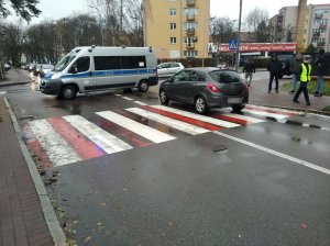 miejsce wypadku drogowego. Na oznakowanym przejściu dla pieszych przed skrzyżowaniem stoi opel corsa. Na skrzyżowaniu stoi policyjny radiowóz blokujący wjazd na ulicę, na której doszło do zdarzenia. Chodnikiem, przy przejściu dla pieszych, idzie policjantka ruchu drogowego