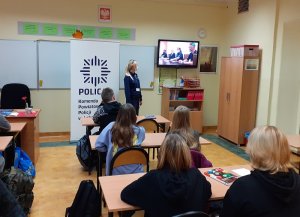 w klasie szkolnej przed tablicą stoi policjantka zwrócona w stronę telewizora, na którym wyświetla się prezentacja. Przed nią w ławkach siedzą uczniowie