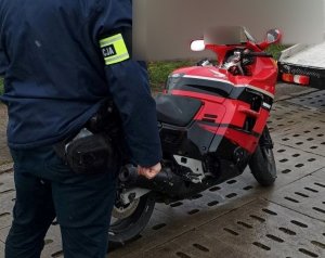 przed garażami stoi nieumundurowany policjant z opaską na ramieniu z napisem policja. Przed nim znajduje się motocykl