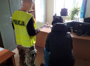 w pokoju przesłuchań na krześle siedzi zatrzymany mężczyzna. Obok niego stoi nieumundurowany policjant w kamizelce z napisem policja