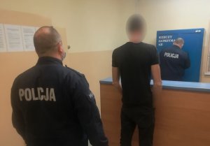 w pomieszczeniu depozytowym stoi zwrócony tyłem do obiektywu zatrzymany mężczyzna. Za nim stoi umundurowany policjant. Przy szafie z depozytem stoi policjant służby dyżurnej