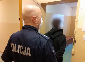 przed otwartymi drzwiami celi stoi skierowany tyłem do obiektywu zatrzymany mężczyzna. Za nim znajduje się policjant służby dyżurnej