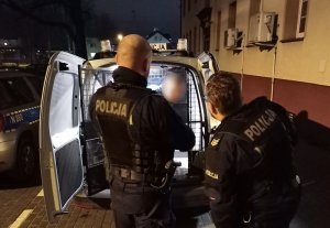 Przed otwartymi tylnymi drzwiami oznakowanego radiowozu stoją dwaj policjanci służby prewencji. W środku znajduje się zatrzymany mężczyzna