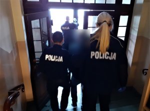 umundurowani policjant i policjantka sprowadzają po schodach zatrzymanego mężczyznę. Przed nimi przy drzwiach wejściowych do komendy stoi policjant służby dyżurnej