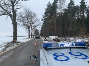 na poboczu drogi stoi skierowany przodem do jezdni biały fiat fiorino. Przód ma zgnieciony. Przed nim po prawej stronie drogi stoi oznakowany policyjny radiowóz. Na drodze stoi strażak