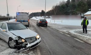 miejsce kolizji. Na zaśnieżonym fragmencie drogi stoi osobowy mercedes z wgniecionym przodem. Za nim stoi policyjny radiowóz. Po prawej stronie drogi stoi policjant ruchu drogowego