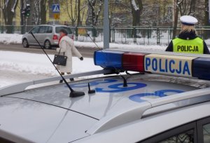 na zaśnieżonym chodniku przed radiowozem stoi policjantka ruchu drogowego, która obserwuje ruch na drodze. Chodnikiem idzie starsza kobieta. Ulicą, w pobliżu przejścia dla pieszych, przejeżdża samochód osobowy