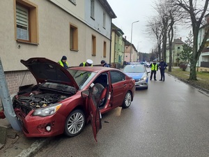 bordowe subaru styka się przodem z latarnią. Latarnia jest wykrzywiona. Subaru ma uniesioną maskę, wgnieciony przód. Drzwi od strony kierowcy są otwarte, widać wystrzeloną poduszkę powietrzną. Przy subaru stoją trzej policjanci ruchu drogowego. W tle widać policyjny radiowóz blokujący drogę oraz dwóch policjantów ruchu drogowego stojących na jezdni