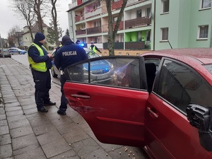 przy bordowym subaru, które ma otwarte drzwi od strony pasażera i uszkodzoną obudowę prawego lusterka oraz zarysowania tylnej prawej części auta stoją policjanci ruchu drogowego. Za subaru stoi policyjny radiowóz