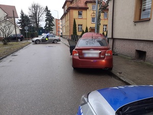 zdjęcie przedstawia bordowe subaru, które zatrzymało się przy latarni i stoi częściowo na jezdni i chodniku. Za subaru stoi oznakowany policyjny radiowóz. Przed subaru w poprzek ulicy stoi nieoznakowany policyjny radiowóz blokujący wjazd na ulicę. W radiowozie siedzi umundurowany policjant