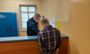 pomieszczenie depozytowe komendy. Przed kontuarem stoi zwrócony tyłem do obiektywu zatrzymany mężczyzna. Przed nim, za kontuarem znajduje się policjant służby patrolowej