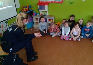 w sali przedszkolnej siedząca na krześle policjantka prowadzi rozmowę z dziećmi siedzącymi w kucki na podłodze