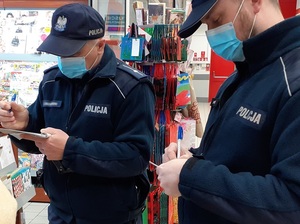 w sklepie znajdują się dwaj umundurowani policjanci w maseczkach ochronnych na twarzach. Jeden z nich wypisuje mandat, drugi wypełnia notatnik