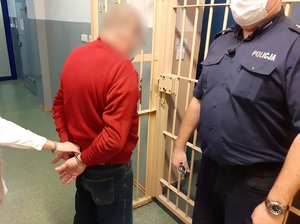 w okratowanym korytarzu pomieszczenia dla osób zatrzymanych stoi mężczyzna z rękami skutymi na plecach kajdankami. Obok niego stoi policjant służby dyżurnej