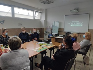 przy stole z seniorkami siedzą dwie umundurowane policjantki z maseczkami ochronnymi na twarzach. Na ścianie na tablicy interaktywnej wyświetla się napis policja ostrzega nie daj się oszukać metoda na wnuczka