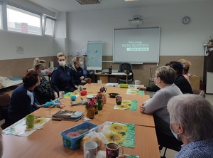 przy stole z seniorkami siedzą dwie umundurowane policjantki z maseczkami ochronnymi na twarzach. Na ścianie na tablicy interaktywnej wyświetla się napis policja ostrzega nie daj się oszukać metoda na wnuczka