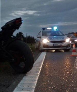 na jezdni stoi oznakowany policyjny radiowóz. Przed nim znajduje się słupek oddzielający drogę od miejsca zdarzenia. Na poboczu stoi motocykl