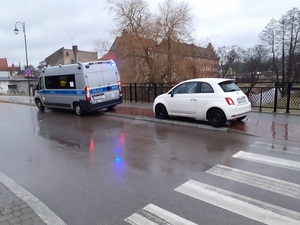 na wydzielonej na chodniku drodze dla rowerów stoi biały fiat 500. Przed nim znajduje się oznakowany policyjny radiowóz z włączonymi sygnałami błyskowymi