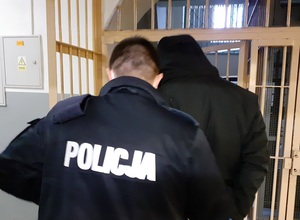 okratowany korytarz pomieszczenia dla osób zatrzymanych. Policjant wyprowadza zatrzymanego mężczyznę