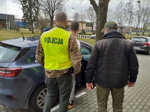 przed nieoznakowanym radiowozem stoją dwaj nieumundurowani policjanci. W radiowozie, na tylne siedzenie siada zatrzymany mężczyzna