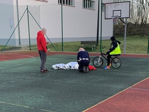 na boisku szkolnym leży postać przykryta kocem termicznym. Przy niej klęczy policjantka. Obok niej stoi mężczyzna z telefonem a na rowerze siedzi kilkunastoletni chłopiec w kurtce policjantki