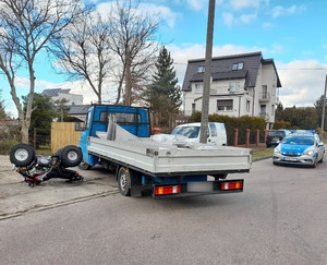 częściowo na jezdni i chodniku, przed wjazdem na teren posesji stoi dostawcze auto. Po jego lewej stronie na chodniku leży przewrócony quad. Na jezdni za pojazdami stoi policyjny radiowóz z włączonymi sygnałami błyskowymi