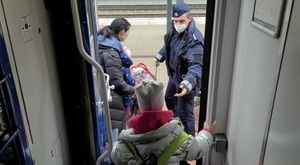 policjant trzymający jedną ręką walizkę wyciąga dłoń do dziecka wychodzącego z pociągu. Obok niego stoi kobieta, która ma na sobie nosidełko z małym dzieckiem