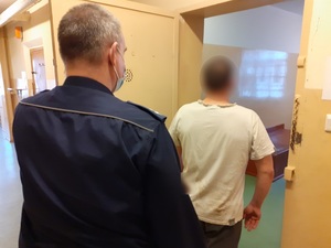 w otwartych drzwiach celi stoi skierowany tyłem do obiektywu mężczyzna. Za nim stoi policjant służby dyżurnej