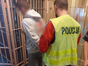 nieumundurowany policjant w kamizelce odblaskowej z napisem policja zakłada zatrzymanemu mężczyźnie kajdanki na ręce trzymane z tyłu