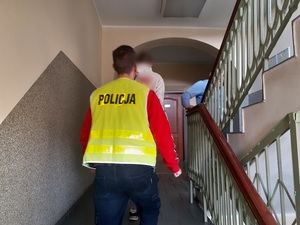 nieumundurowany policjant w kamizelce odblaskowej z napisem policja prowadzi schodami w górę zatrzymanego mężczyznę