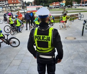 na placu stoi policjant ruchu drogowego. Przed nim znajdują się dzieci z rowerami. Dzieci mają na sobie kamizelki odblaskowe