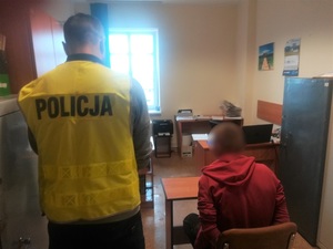 na krześle w pokoju przesłuchań siedzi mężczyzna. Po jego lewej stronie stoi policjant w kamizelce odblaskowej z napisem policja