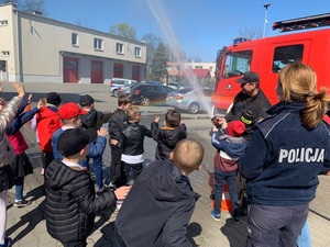 na terenie komendy straży pożarnej strażak prezentuje dzieciom wąż strażacki, z którego leci woda. Obok stoi umundurowana policjantka