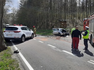 po obu stronach prostej drogi znajdują się dwa osobowe samochody uczestniczące w wypadku. Po lewej stronie znajduje się ustawiona w poprzek biała kia sportage, na poboczu z prawej strony drogi srebrny vw golf z licznymi uszkodzeniami w postaci wgnieceń karoserii. Na miejscu są strażacy i policjant ruchu drogowego oraz wóz strażacki