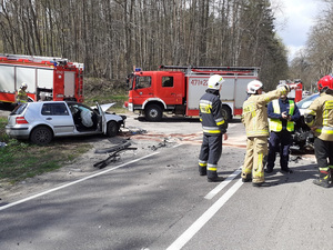 po obu stronach prostej drogi znajdują się dwa osobowe samochody uczestniczące w wypadku. Po prawej stronie znajduje się ustawiona w poprzek biała kia sportage z wgniecionym przodem, na poboczu z lewej strony drogi srebrny vw golf z licznymi uszkodzeniami w postaci wgnieceń karoserii. Na miejscu są strażacy i policjant ruchu drogowego oraz wozy strażackie i karetka pogotowia. Strażak wskazuje coś ręką policjantowi