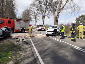 miejsce wypadku. Na prawej stronie jezdni znajduje się ustawiona w poprzek biała kia sportage z wgniecionym przodem. Przed nią stoją strażacy i policjant ruchu drogowego. Za nią znajduje się karetka pogotowia. Z lewej strony drogi, na poboczu znajduje się srebrny osobowy samochodów z wystrzeloną poduszką powietrzną i podniesioną maską. Przed nim stoi wóz strażacki. Na jezdni znajdują się odłamki z samochodów