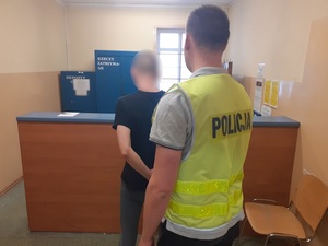 w pomieszczeniu dla osób zatrzymanych stoi zwrócony tyłem do obiektywu mężczyzna z rękoma skutymi kajdankami. Za nim stoi nieumundurowany policjant w kamizelce z napisem policja