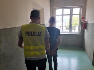 nieumundurowany policjant w kamizelce odblaskowej z napisem policja prowadzi korytarzem zatrzymanego mężczyznę, który ma ręce skute kajdankami