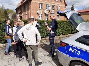 przy otwartych drzwiach bagażnika radiowozu stoi policjant ruchu drogowego, który prezentuje dzieciom policyjną pałkę służbową