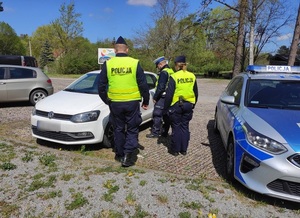 troje umundurowanych policjantów w kamizelkach odblaskowych stoi przy białym osobowym volkswagenie. Jeden z policjantów nachyla się w stronę kierowcy