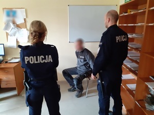 w pomieszczeniu komendy umundurowani policjant i policjantka stoją przed siedzącym na krześle mężczyzną, który ma ręce skute kajdankami