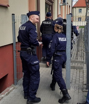 dwaj umundurowani policjanci prowadzą w kierunku wejścia do komendy zatrzymanego mężczyznę. Za nimi idzie policjantka. Obok niej stoi policjant