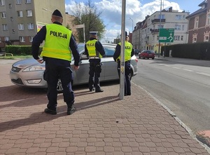 troje umundurowanych policjantów stoi przed zaparkowanym na chodniku srebrnym osobowym kombi. Jeden z policjantów, funkcjonariusz ruchu drogowego, znajduje się przy drzwiach od strony kierowcy