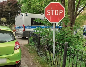 na chodniku wzdłuż jezdni stoi policyjny radiowóz. Przed chodnikiem, prostopadle do jezdni  obok znaku drogowego &quot;stop&quot; stoi zielony osobowy samochód