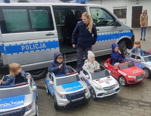 przed oznakowanym radiowozem stoi policjantka. Przed nią w zabawkowych samochodach policji, pogotowia ratunkowego i straży pożarnej siedzą kilkuletnie dzieci