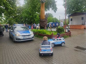 festyn. Na chodniku stoi oznakowany policyjny radiowóz. Przed nim na zabawkowych samochodzikach - radiowozach jeżdżą dzieci