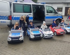 przed oznakowanym policyjnym radiowozem, w zabawkowych samochodach policji, pogotowia ratunkowego i straży pożarnej siedzą kilkuletnie dzieci. Policjantka nachyla się w kierunku dziewczynki siedzącej w zabawkowym radiowozie