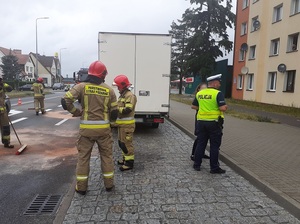 na drodze strażacy uprzątają jezdnię po kolizji. Za uczestniczącym w kolizji białym iveco stoi policjant, który rozmawia z kobietą